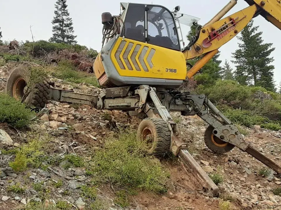 A yellow walking excavator braced on its legs across a steep, rocky slope among pines, a drill mast on its boom