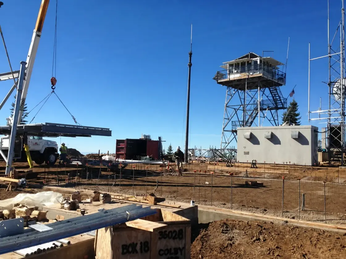 A crane lifting steel members beside a tall steel lookout tower on a mountain summit