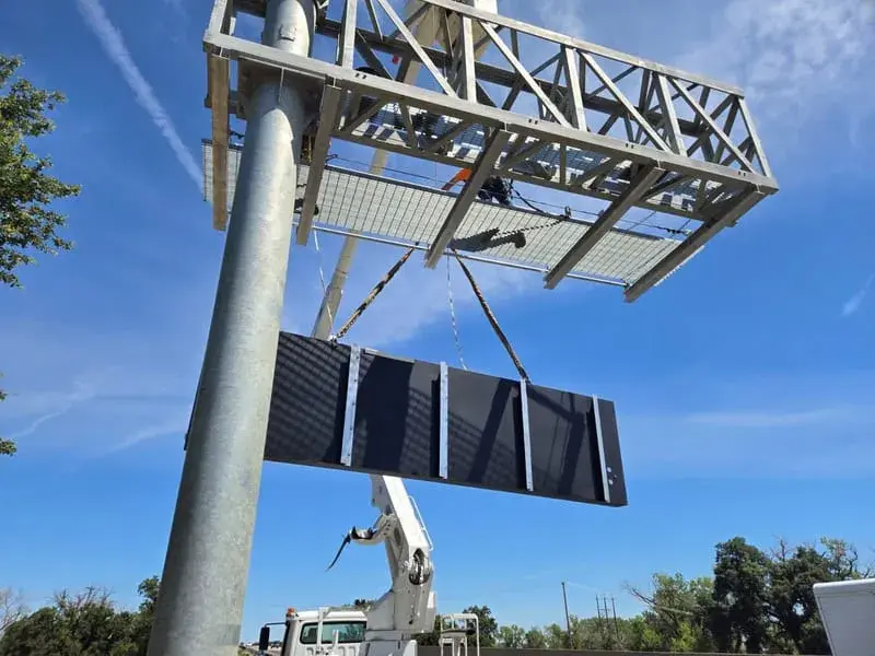 A crane lifting a steel overhead sign truss up to a mounting post against a blue sky