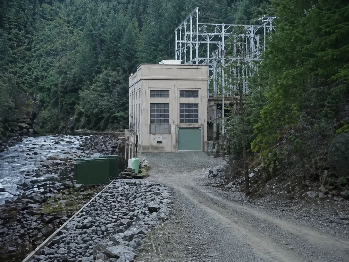 A small concrete powerhouse beside a river with an outdoor switch structure behind it