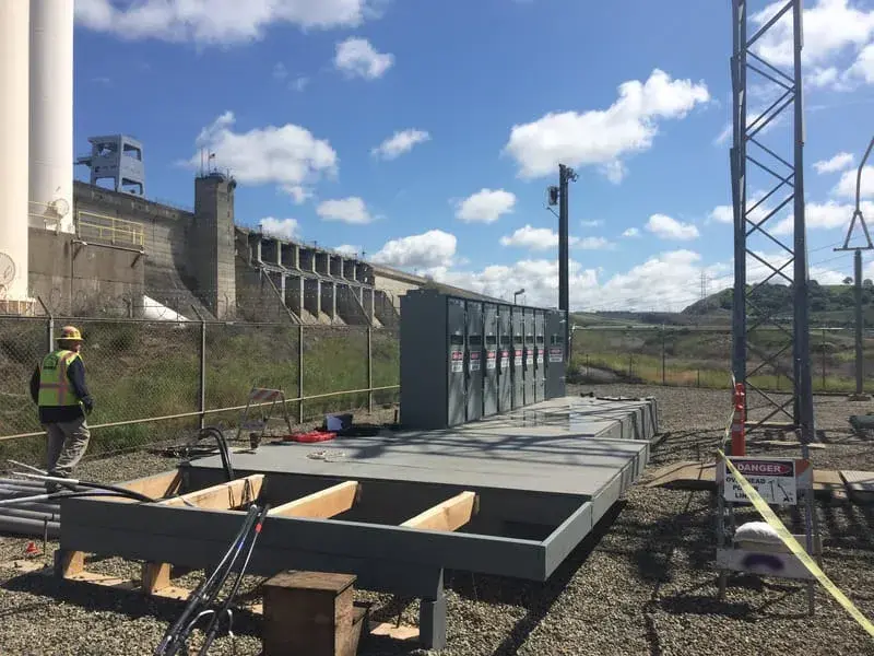 New electrical enclosures on a concrete pad with the spillway gates of a large dam rising behind