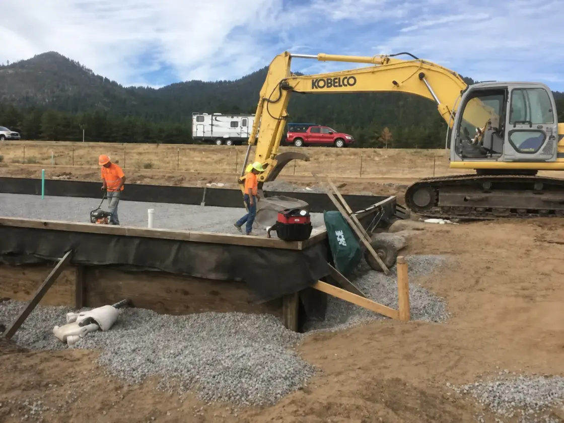 A crew finishing a new concrete slab within timber formwork, with an excavator alongside