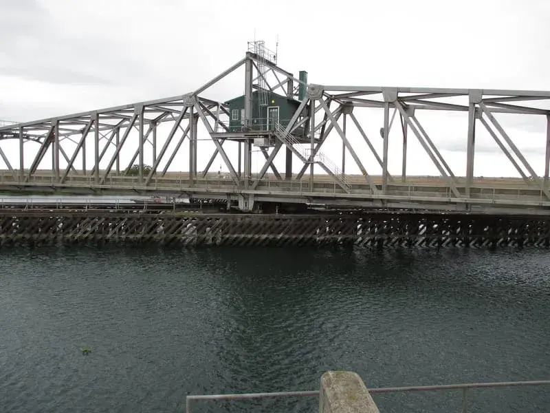A long steel swing bridge over water, seen from the levee with timber pilings along the shore