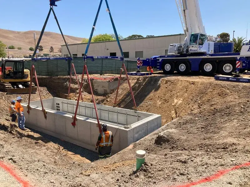 A crane lowering a large precast concrete vault on four slings into an open excavation while a crew guides it