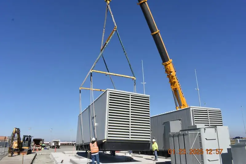 A crane lifting a large radiator-fronted generator set above a completed concrete pad