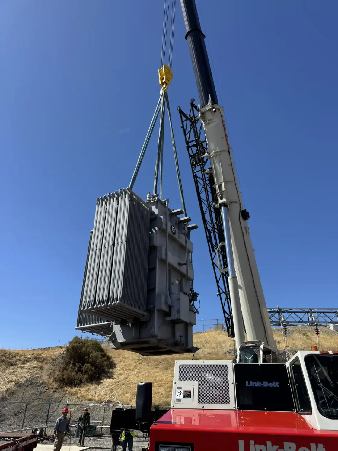 A single-phase power transformer suspended from crane rigging above a substation yard, dry hills behind