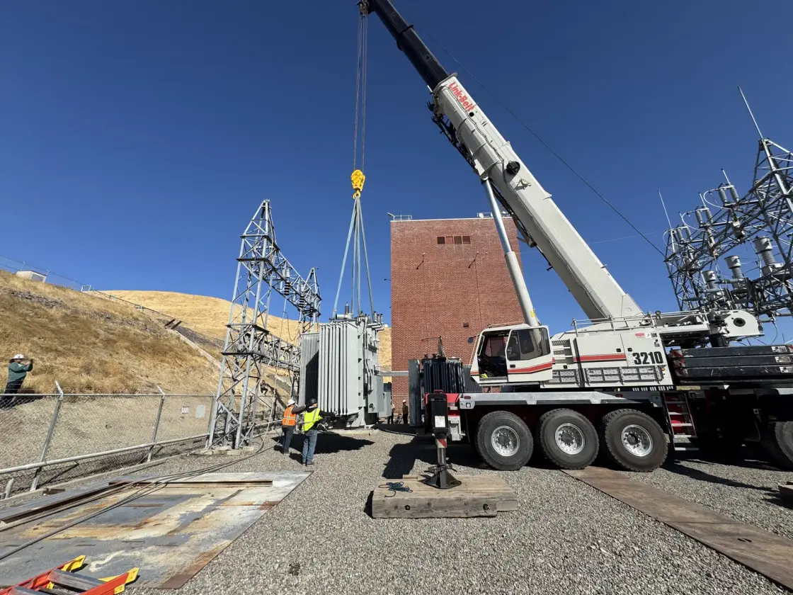 A wide view of a pumping plant with a mobile crane rigged beside the switchyard and a crew working at the base