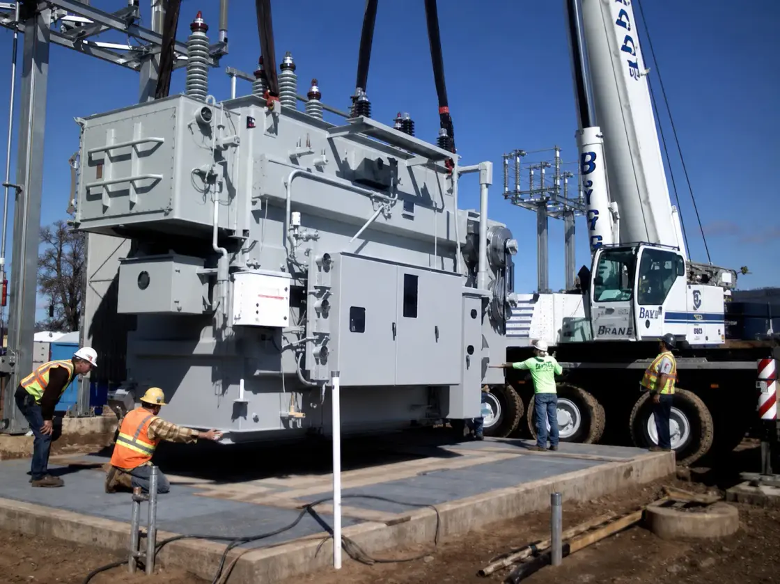 Workers guiding a large power transformer down onto a concrete pad between two cranes