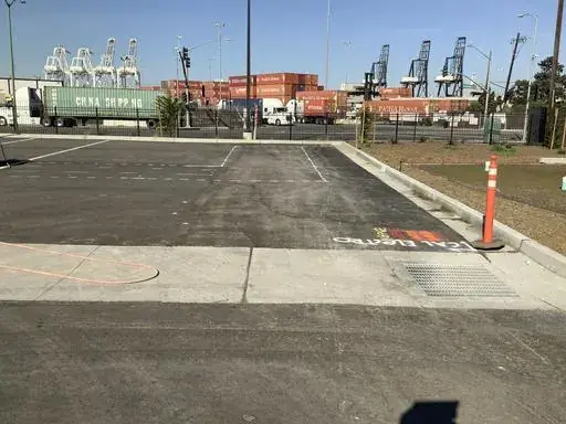 A marked saw-cut line crossing a paved lot toward a fence, stacked shipping containers and ship-to-shore cranes beyond
