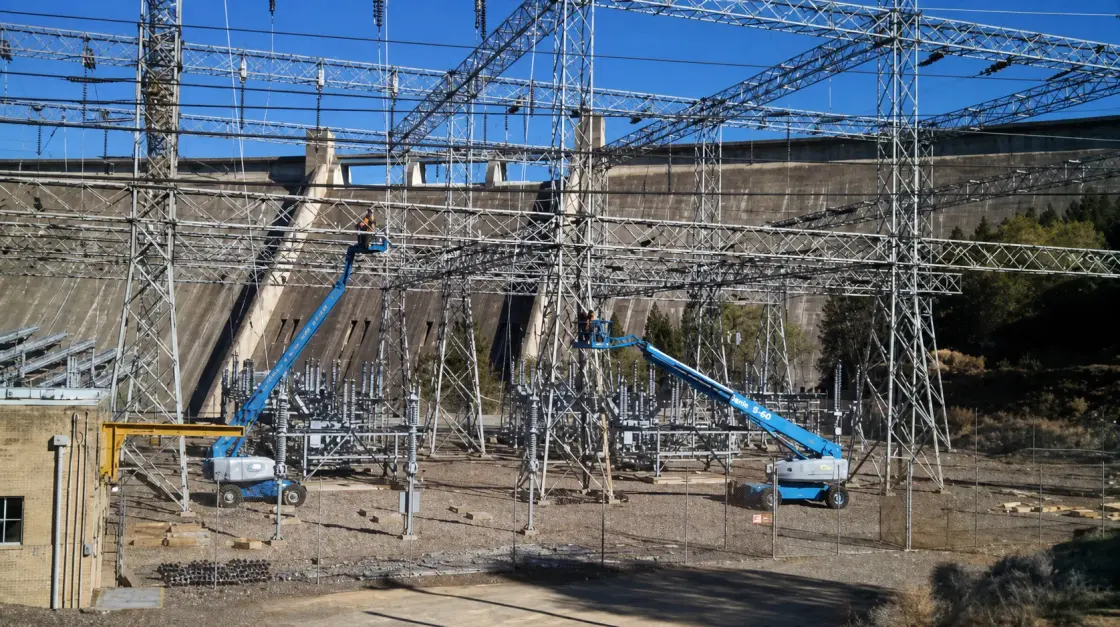 A lift working beneath the steel bus structures of a switchyard, with the face of a large concrete dam behind