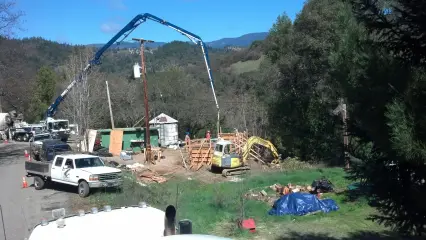 A concrete pump truck with its boom extended over a hillside construction site, with a storage tank behind