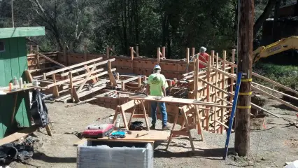 Timber formwork braced into position around a foundation, with a crew working at a cut table alongside