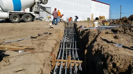 A long trench with reinforcing steel and conduit laid in, running along the side of a building