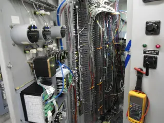 An open control cabinet with dense wire bundles, labelled control switches and a digital multimeter hanging at the right