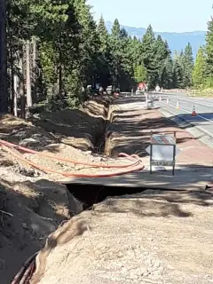 An open trench with conduit alongside a highway lined with pines, traffic cones set along the lane