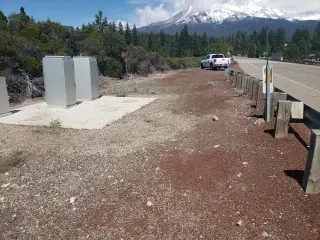 Two grey equipment cabinets on a concrete pad beside a guard-railed highway, with a snow-capped mountain beyond the trees