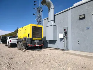 A yellow towable generator parked beside a grey equipment building, a communications tower rising behind it