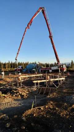 A concrete pump boom extended over a foundation excavation on a wooded mountaintop at dusk