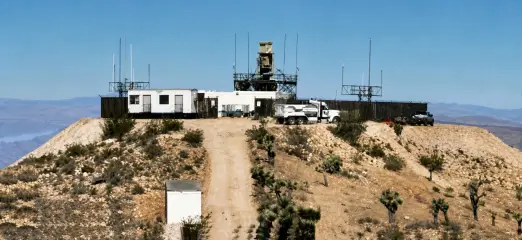A fenced radar and communications compound on a bare desert hilltop, with masts and a radome