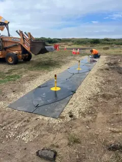 A yellow airfield light fitting set into a black mat on a gravel runway shoulder, with a loader working behind