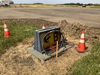 A black airfield sign base with cabling installed, sitting on a concrete pad beside the runway and marked with cones