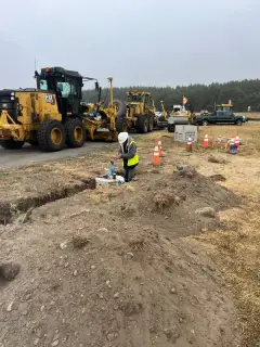 A grader and a crew working along an open trench beside a runway