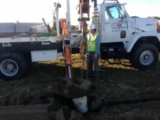 A worker beside a boom truck fitted with an auger, setting a pole in freshly turned ground