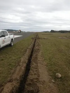 A long narrow trench cut through grass alongside a runway, with a white truck parked at the far end