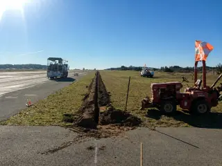 A tracked trencher cutting a trench along the edge of a runway, with marker flags alongside