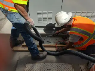 A worker in a hard hat and vest drilling at the base of new switchgear while a second worker holds a vacuum hose to the hole