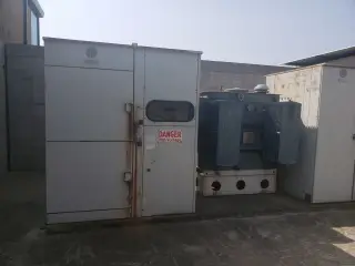 A weathered outdoor switchgear enclosure and transformer standing in a walled yard, with a high-voltage warning sign