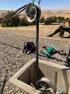 Cable running over a pulling sheave down into a concrete pull box set in gravel, with dry hills in the distance