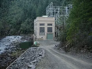A small concrete powerhouse beside a river with an outdoor switch structure behind it