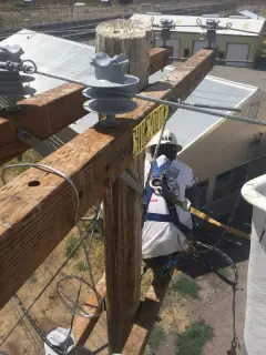 A worker in fall protection reaching along a new wooden crossarm fitted with insulators