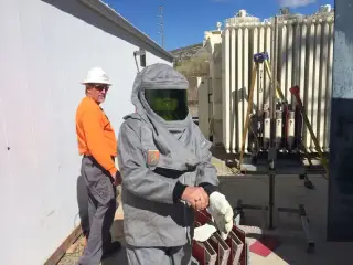 Two workers at an open electrical cabinet, one in a full silver arc flash suit and hood, the other in high-visibility clothing