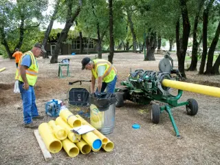 Two workers at a fusion machine beside stacked lengths of yellow gas pipe under oak trees