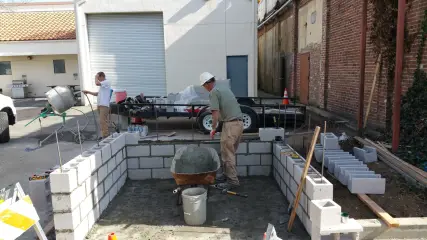 Two workers laying concrete block for a low enclosure wall beside a mixer and a stack of block