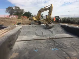 An excavator spreading gravel across a membrane-lined basin at a highway rest area site