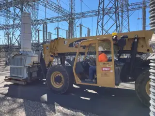 A telehandler carrying a station service transformer past crew members inside a substation yard