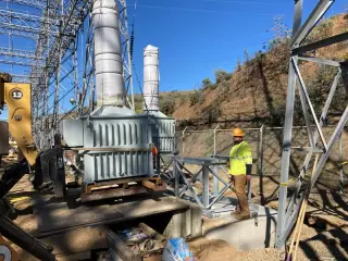 Workers guiding a station service transformer into position on a pad at the base of a steep hillside