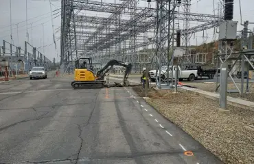 An excavator working along a paved road that runs through a substation, with steel structures on both sides