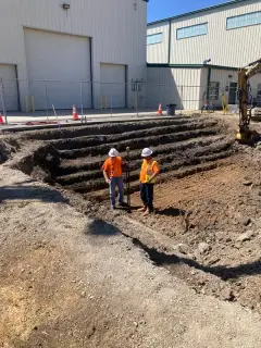 Two workers standing in a stepped excavation beside a large industrial building