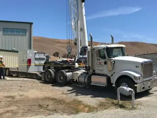 A flatbed truck carrying electrical equipment parked beside a building, with a crane working in the background