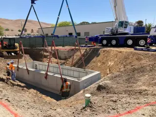 A crane lowering a large precast concrete vault on four slings into an open excavation while a crew guides it