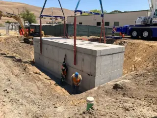 A precast concrete vault set in an excavation with a worker connecting conduit at one end