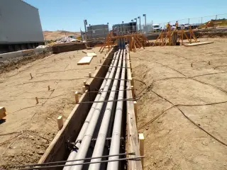 Multiple parallel grey conduits laid in a long open trench across a graded site