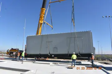 A crane lowering a long grey walk-in electrical enclosure onto a new concrete pad