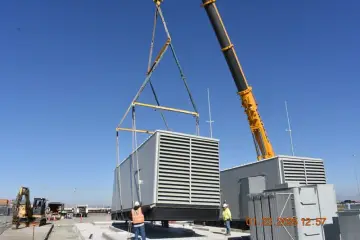 A crane lifting a large radiator-fronted generator set above a completed concrete pad