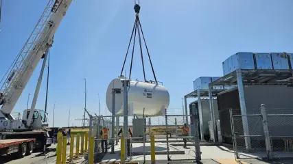 A crane lowering a horizontal cylindrical fuel tank beside new electrical equipment