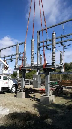 Electrical equipment slung on red rigging beneath a crane hook, being lowered between steel structures